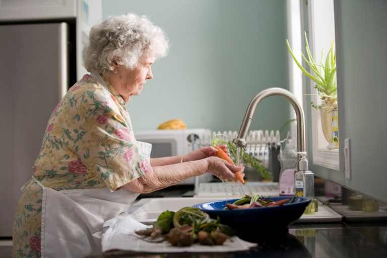 Elderly women washing carrots in kitchen sink. A bowl of other vegetables sits on the counter nearby.