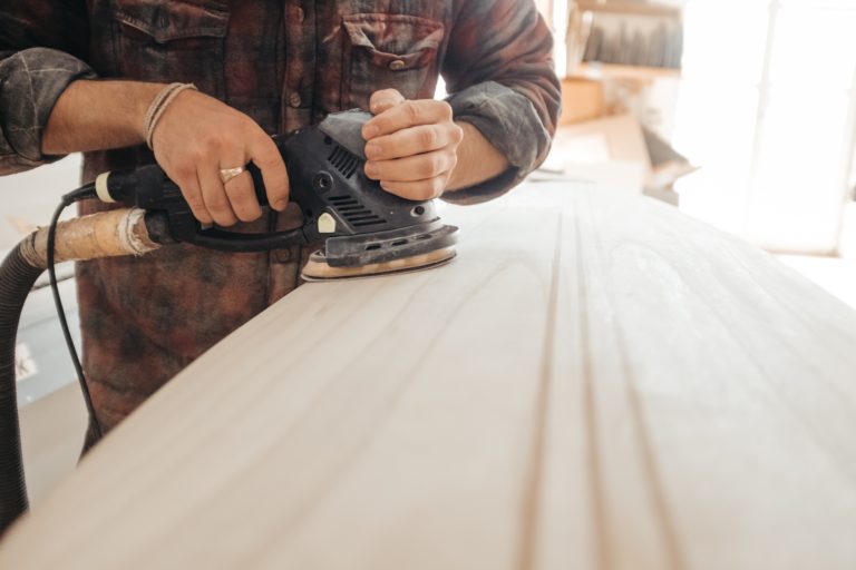 A person using an electric dander to smooth a wide plank of wood. Their clothes are covered in wood shavings.